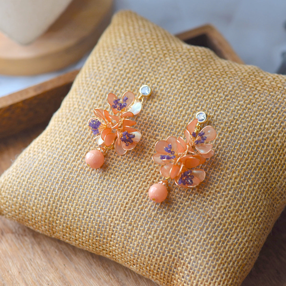 Apricot Fleur apricot-orange resin floral earrings with lilac stamens and peach drops on jute cushion
