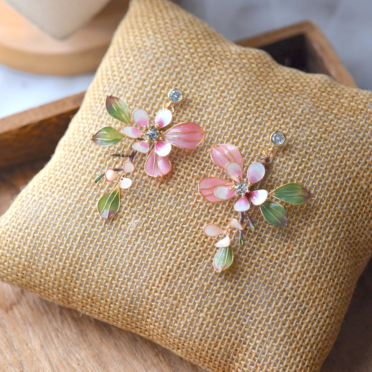 Florentine blush pink resin floral earrings with olive green leaves and zirconia centres on a jute cushion
