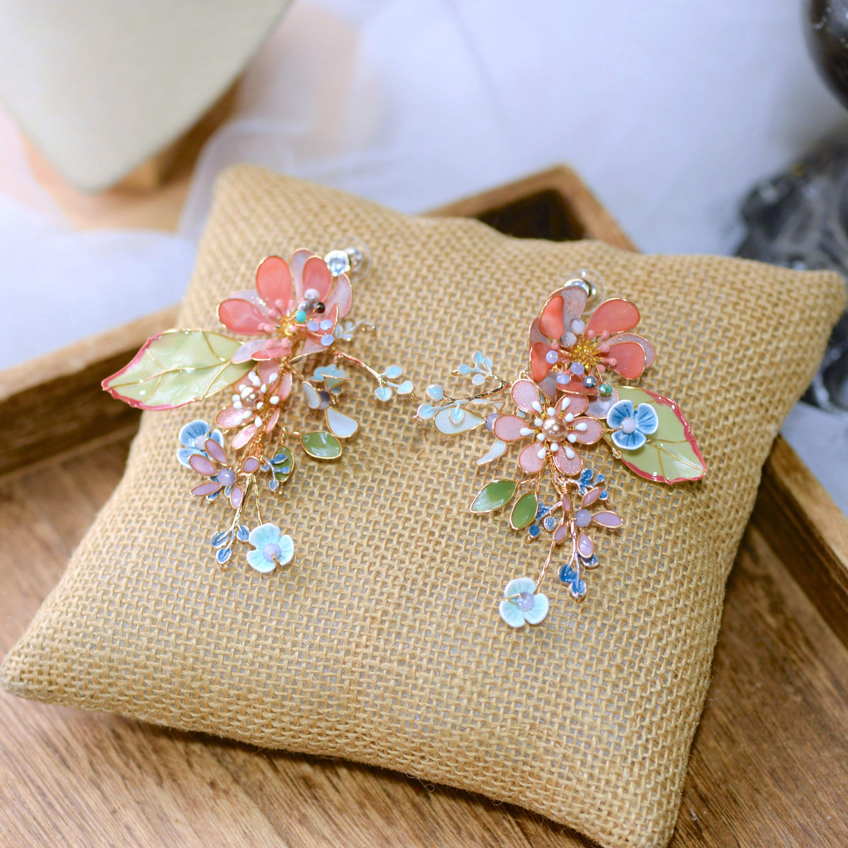 Trina coral and green resin floral earrings with golden accents laid on a jute cushion
