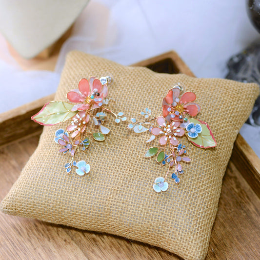 Trina coral and green resin floral earrings with golden accents laid on a jute cushion
